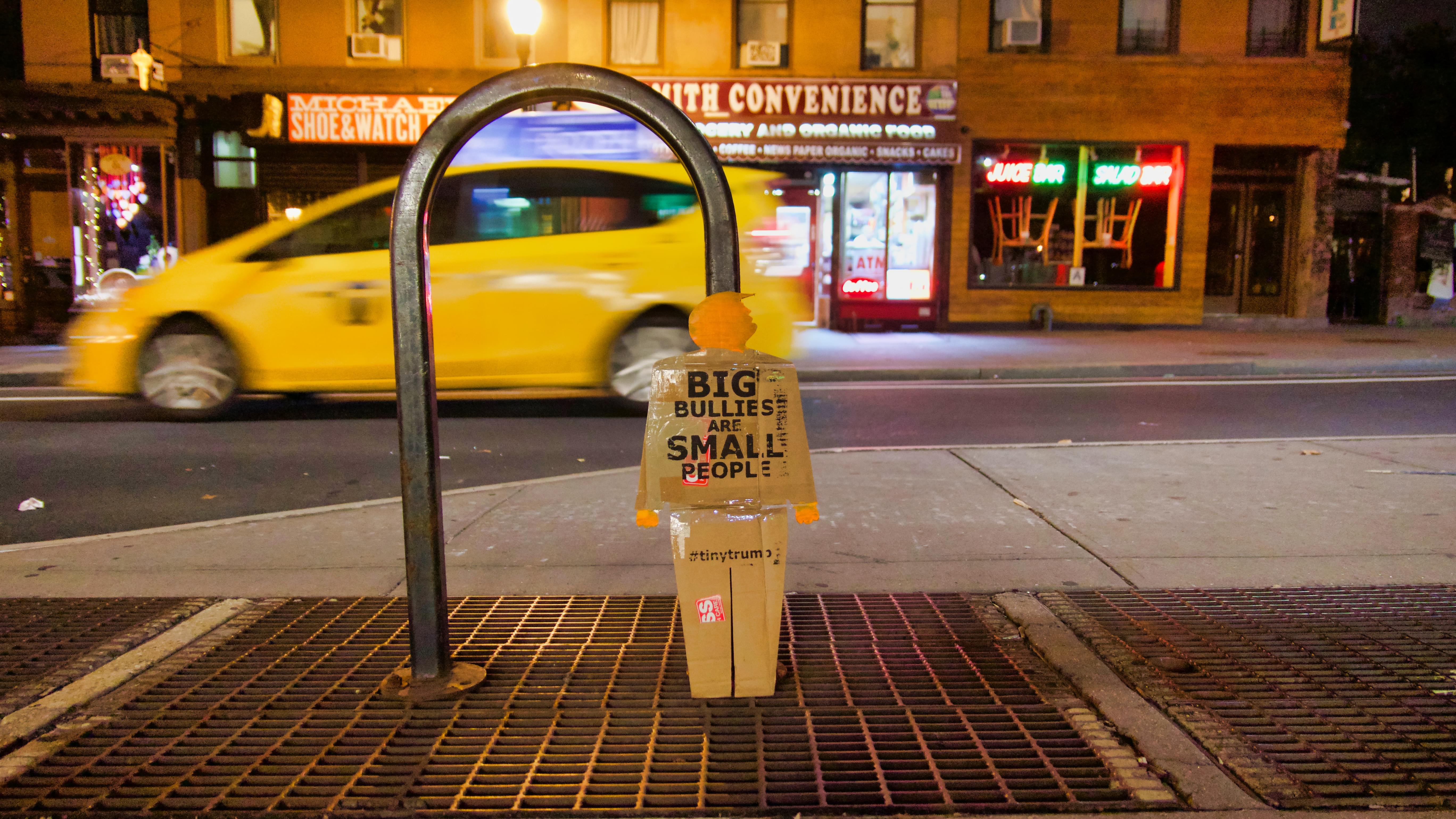 A two-foot-tall cardboard cutout tiny trump protest sign (in the image of a full-bodied trump) leaning against a bike rack in the street in Brooklyn, New York. It is nightime and a yellow cab is blurred in the background as it whizes by.