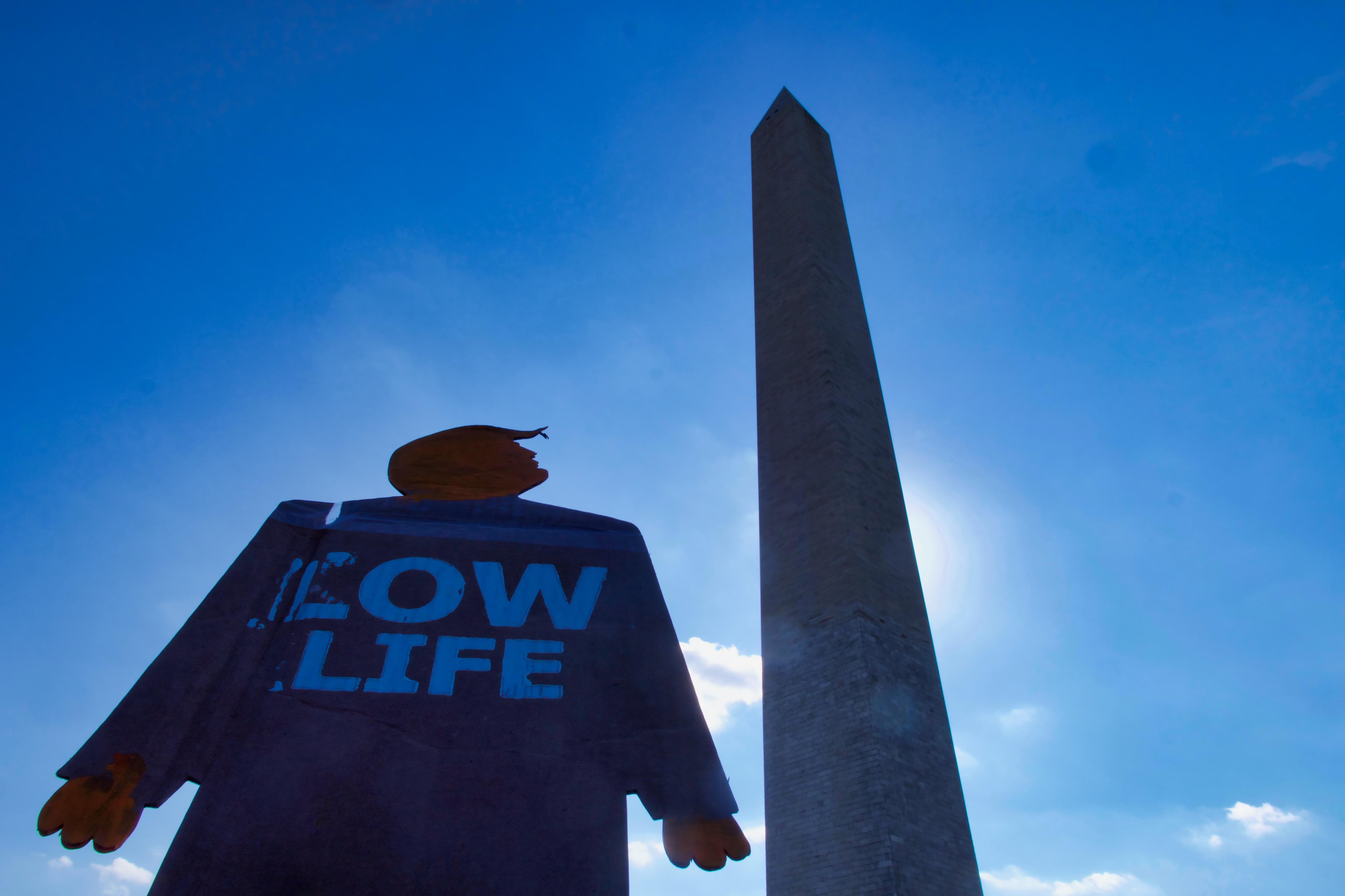 A two-foot-tall cardboard cutout tiny trump protest sign with the slogan Low Life looking up at the Washington Monument in Washington, D.C. Photograph angle is pointed up sharply so only the tiny trump, monument, and sky are visible