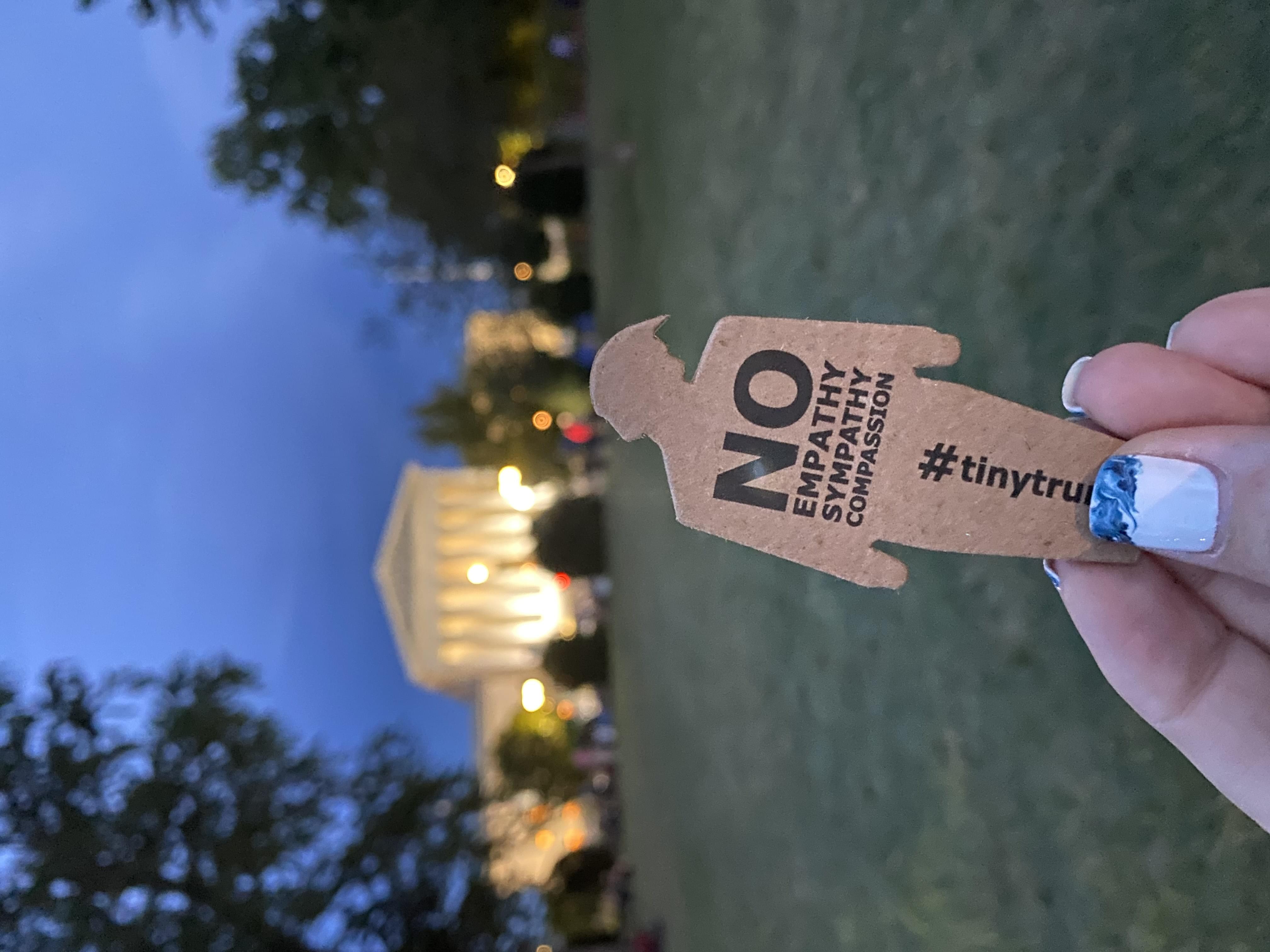 a photograph of someone holding a tiny trump with the slogan 'No Empathy, No Sympathy, No Compassion' in front of the White House