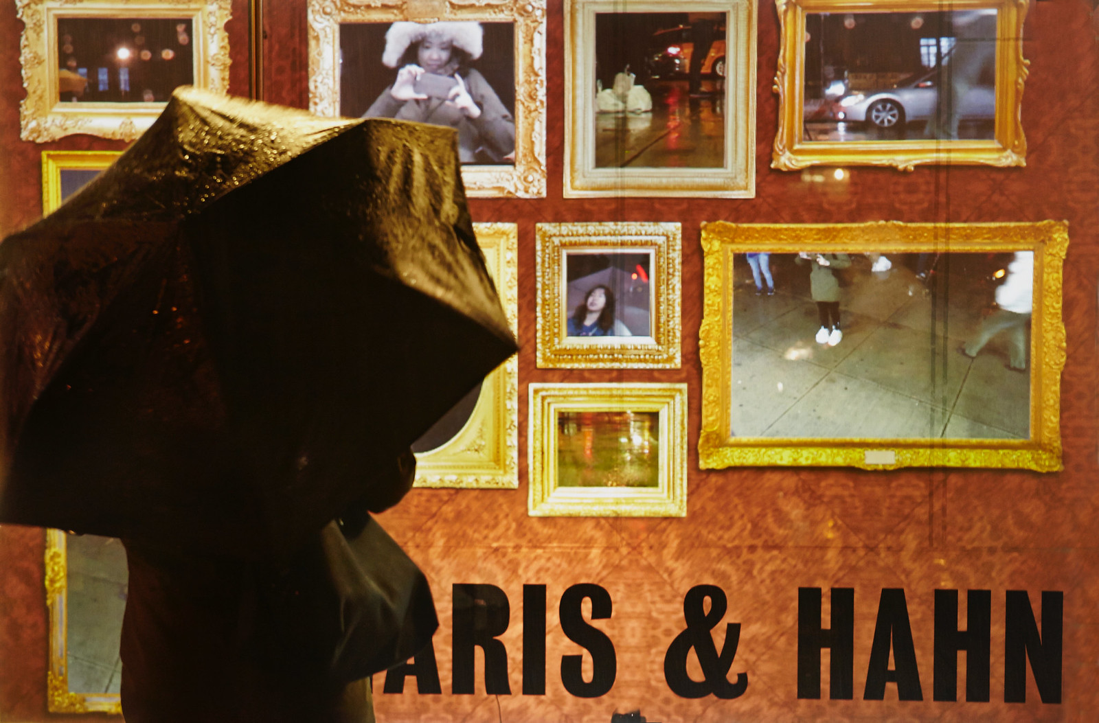 Photograph of someone with an umbrella, outside in the rain, looking at a gallery storefront which shows a projection of nearly a dozen golden-framed video feeds, some of which show different facets of the viewer (her face, shoes, torso), some of which show facets of the urban landscape in the background (sidewalk, street, cars, etc.).