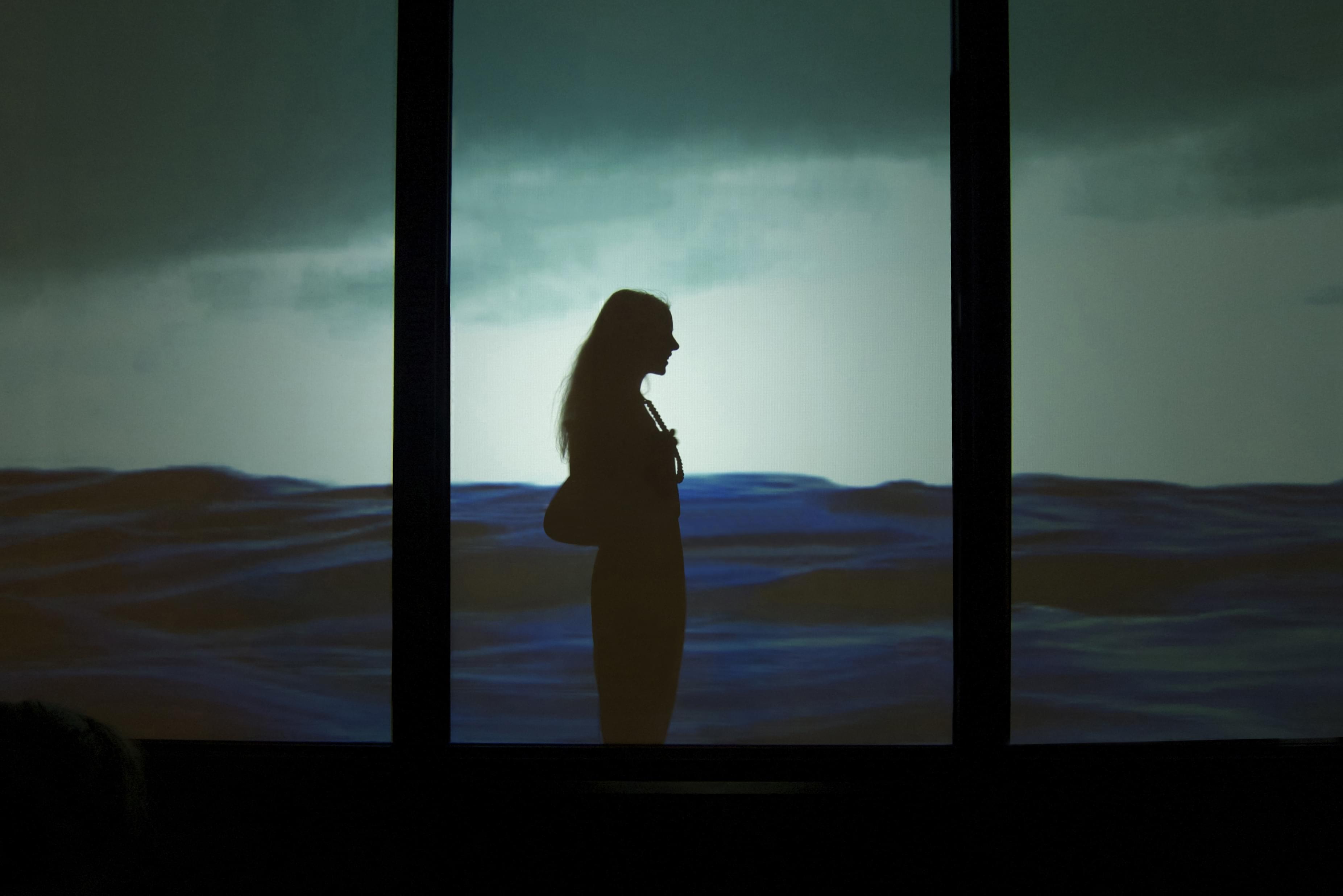 Nighttime photograph showing a urban storefront showing a video of a contiguous body of water projected across 4 of its windows; lower half of the project is dark blue water, the upper half is light blue sky