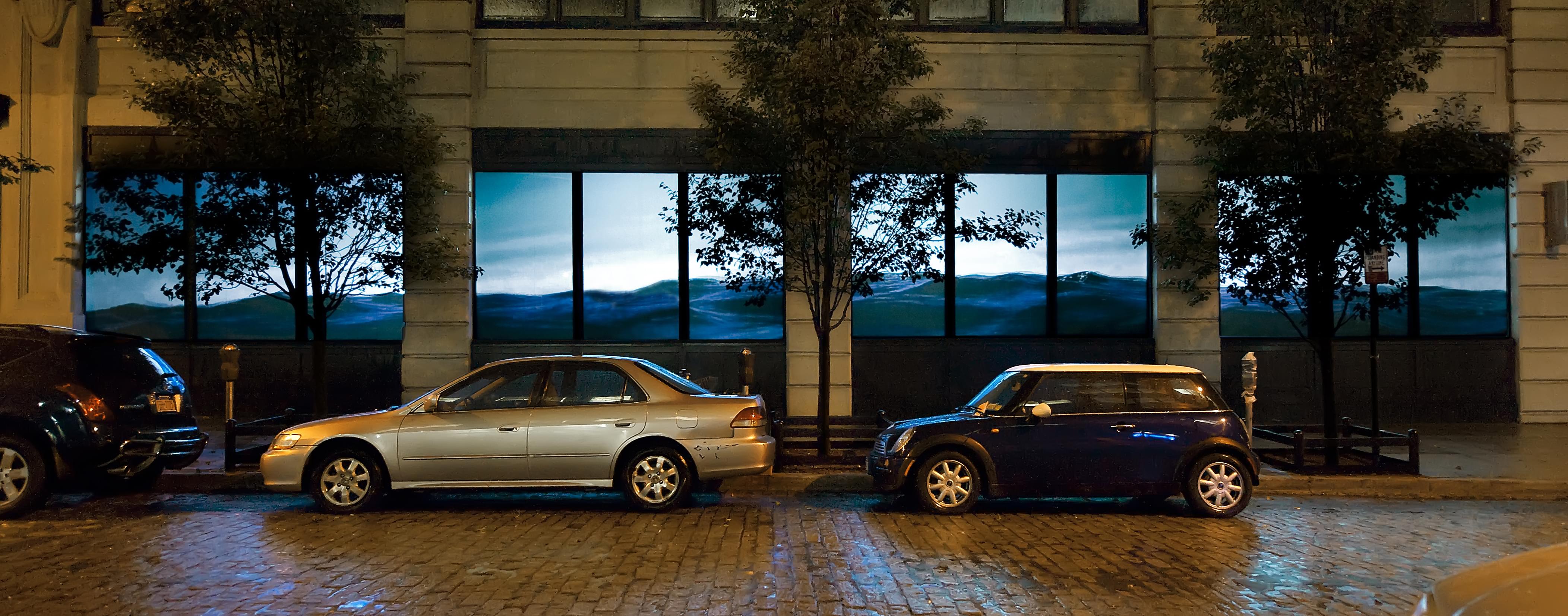 Nighttime photograph showing a urban storefront showing a video of a contiguous body of water projected across 4 of its windows; lower half of the project is dark blue water, the upper half is light blue sky