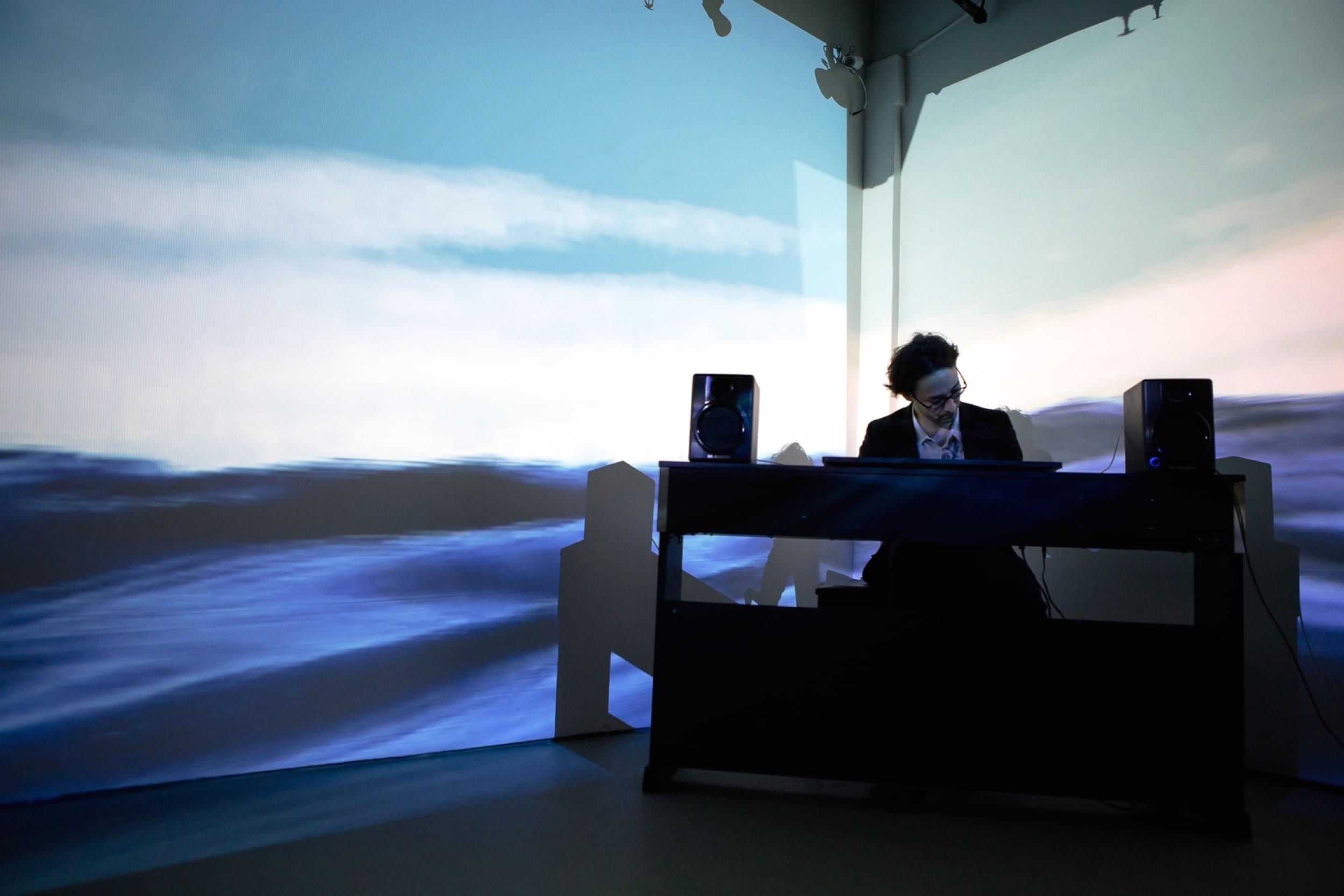 Photograph of the artist, Eric Corriel, swaying while playing piano in the corner of a gallery, which is mostly dark. Projected onto the two walls that form the corner is his piece, Water Will Be Here, which I'm sure you've guessed by now, consists of a video of a body of water.