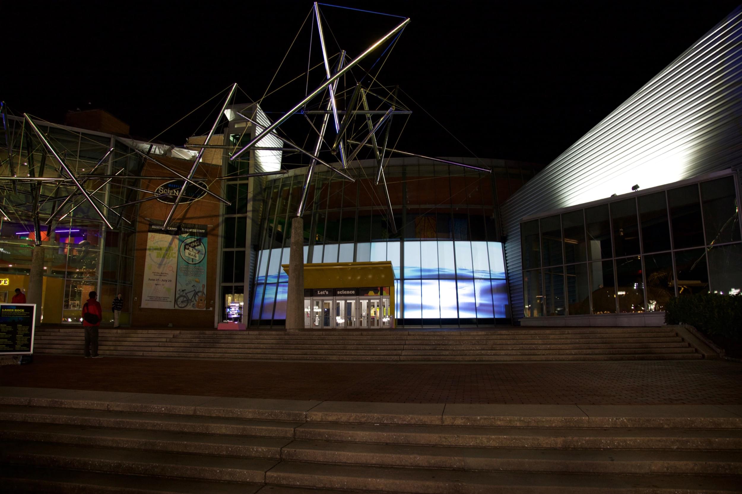 Overhead shot of a gala event&mdash;dozens of elegantly decorated tables with white table cloths and well-dressed people sitting at them. Behind them, on a long white wall, a video of a body of water is projected