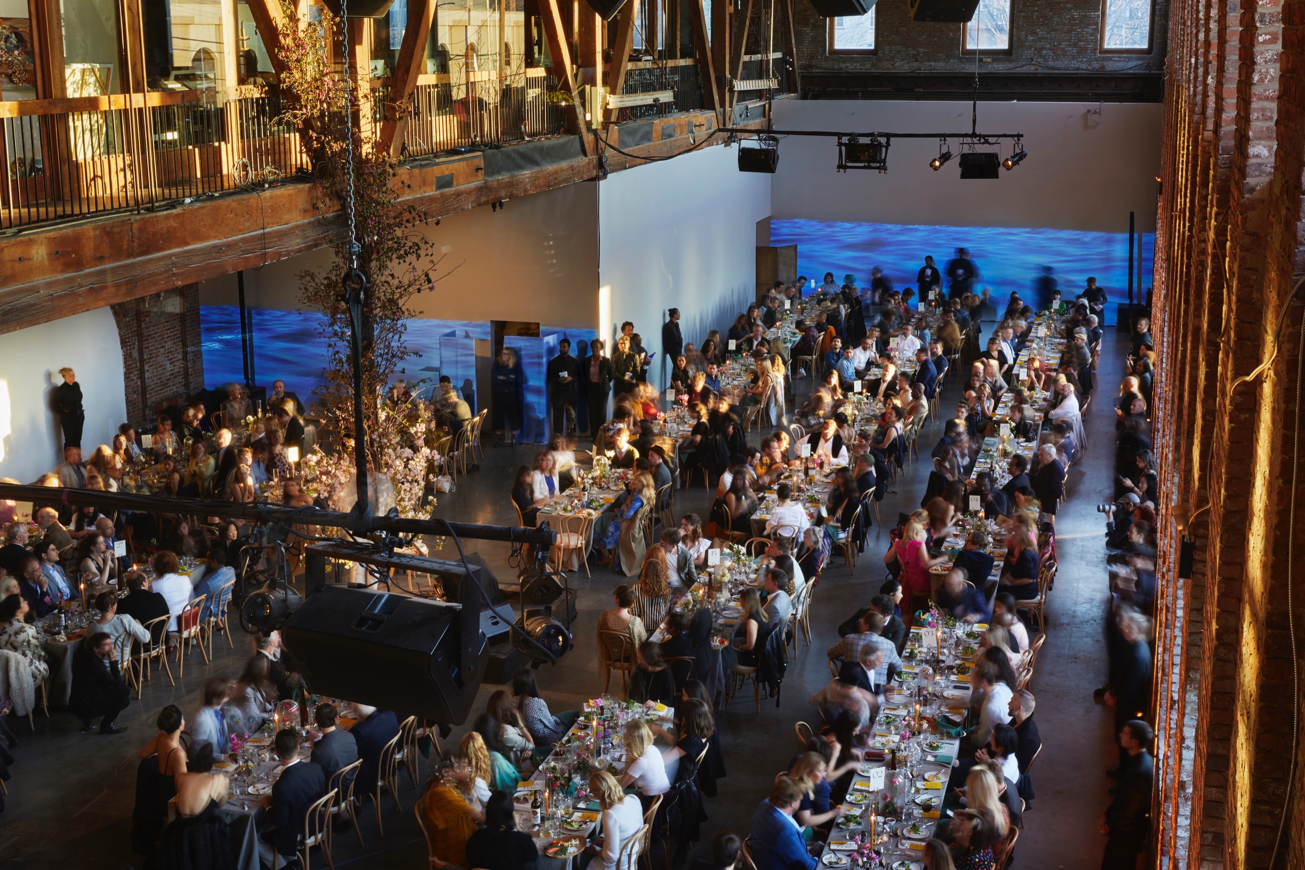 Overhead shot of a gala event&mdash;dozens of elegantly decorated tables with white table cloths and well-dressed people sitting at them. Behind them, on a long white wall, a video of a body of water is projected