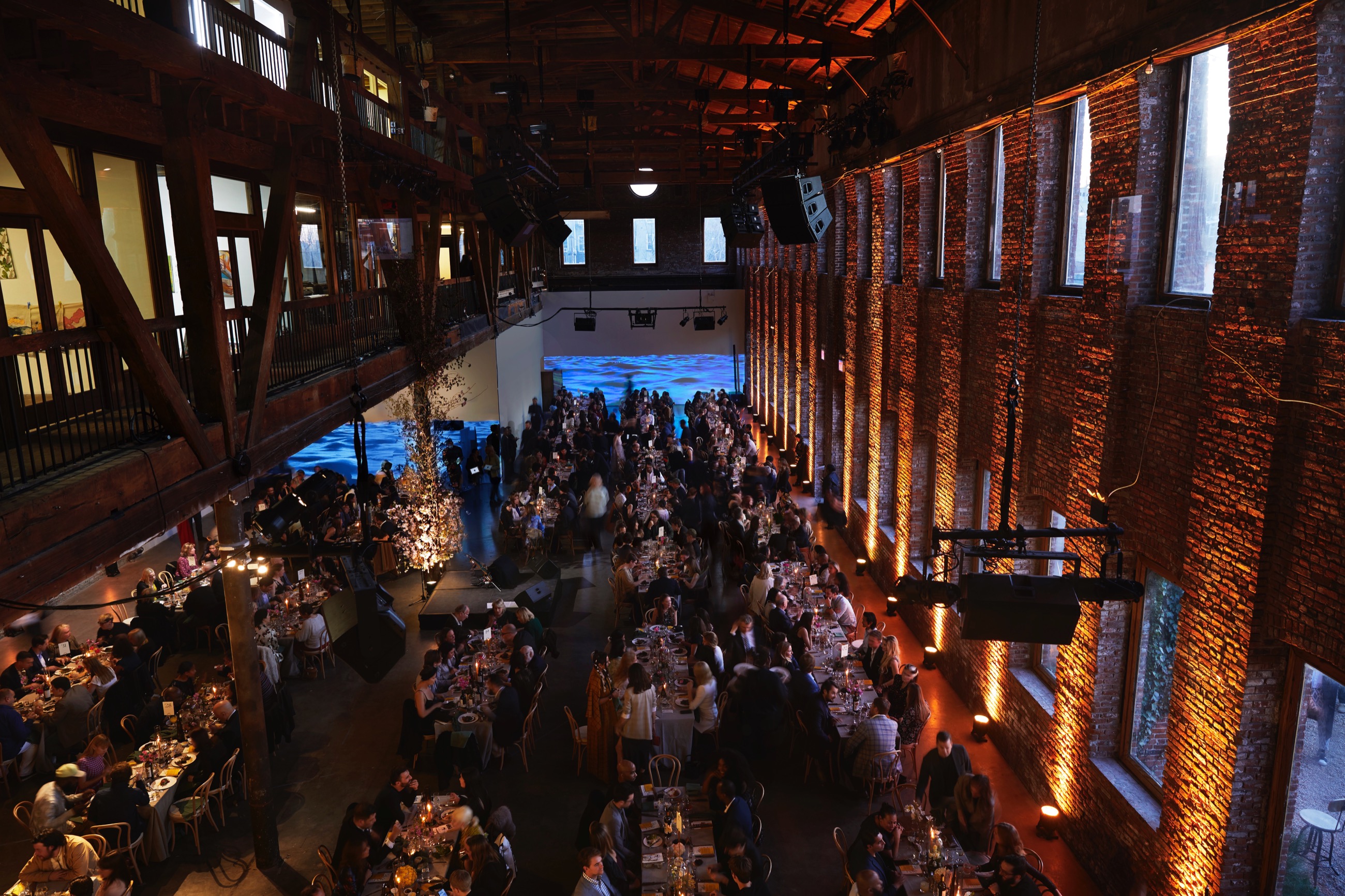 Overhead shot of a gala event, over elegantly decorated tables with well-dressed people seated at them. Behind them, on a long white wall, a video of a body of water is projected 