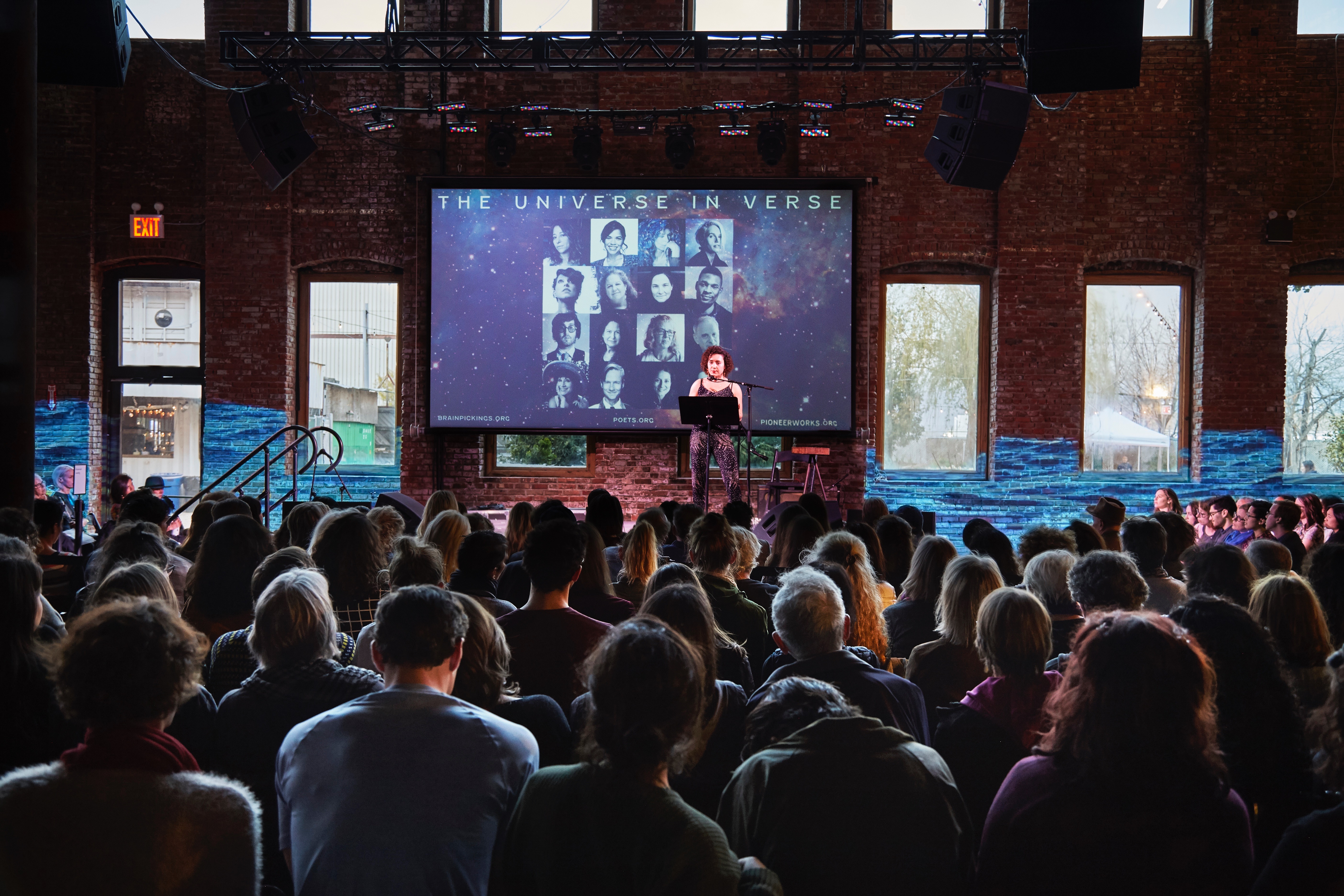 Photograph of an event in which approximately a hundred people are seated&mdash;we see the backs of their heads. In the background is a woman on stage, reading (a poem). Behind her is a screen with text that reads 'The Universe In Verse,' which is flanked on both sides by a projection of video of water on a brick wall.