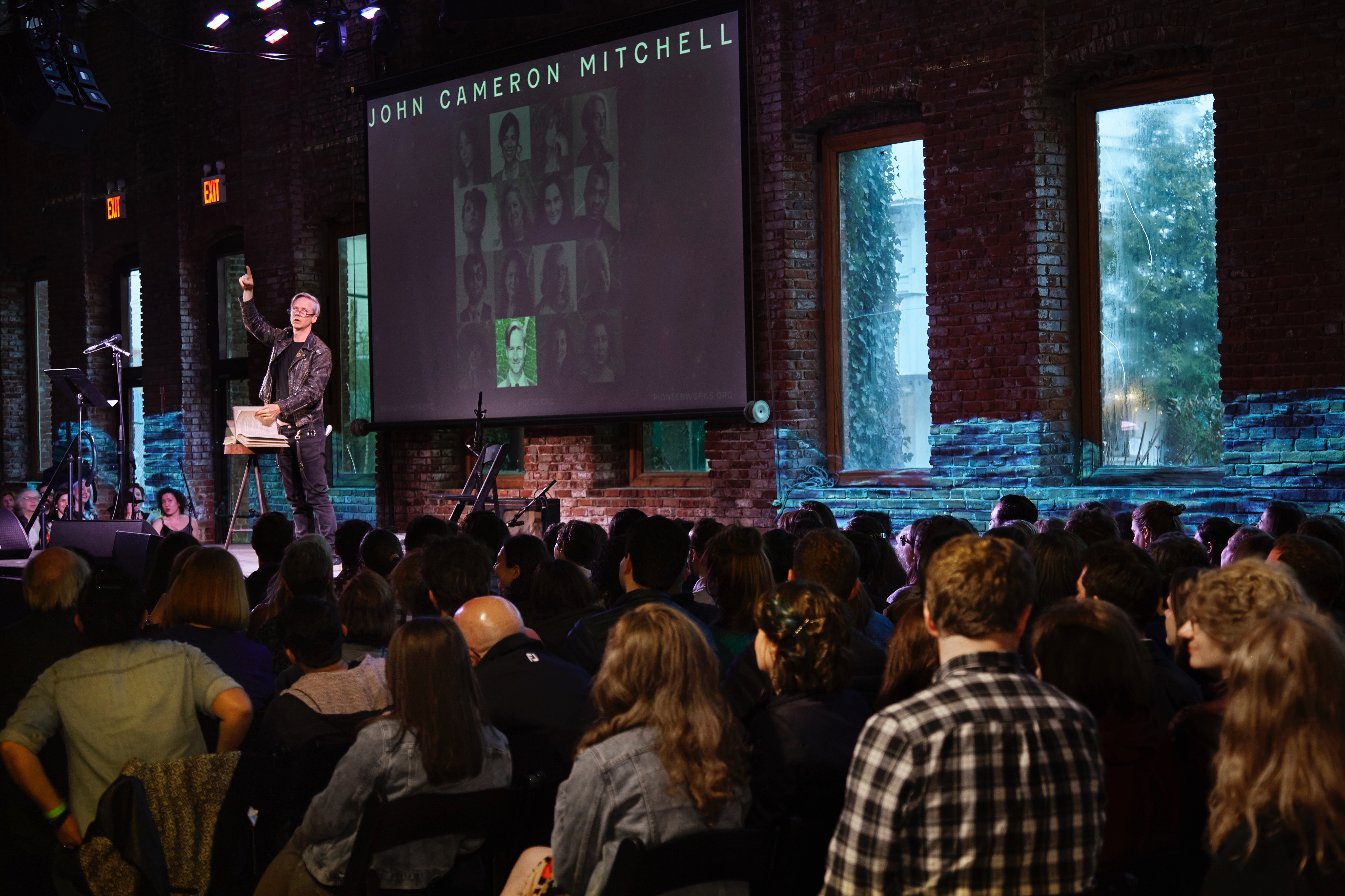 John Cameron Mitchell gestures with a finger raised to the sky as he reads a poem to an audience. A video of a body of water is projected onto the interior brick wall behind him.
