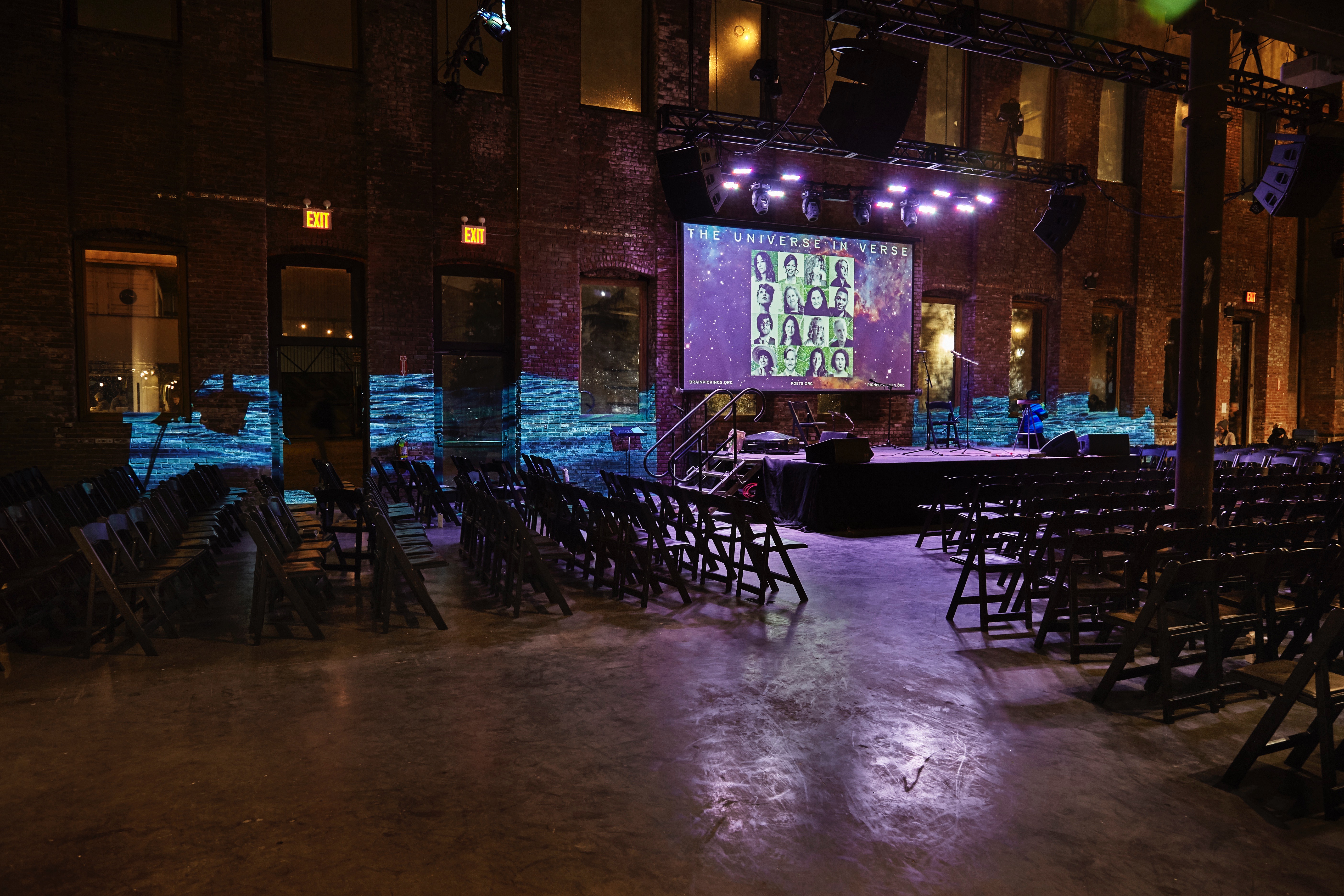 Photograph of a large empty room with over a hundred empty folding chairs. A small stage is in the center and above it, a screen indicates the name of the event: 'The Universe In Verse.' A video of a body of water is projected on the brick wall in the background.