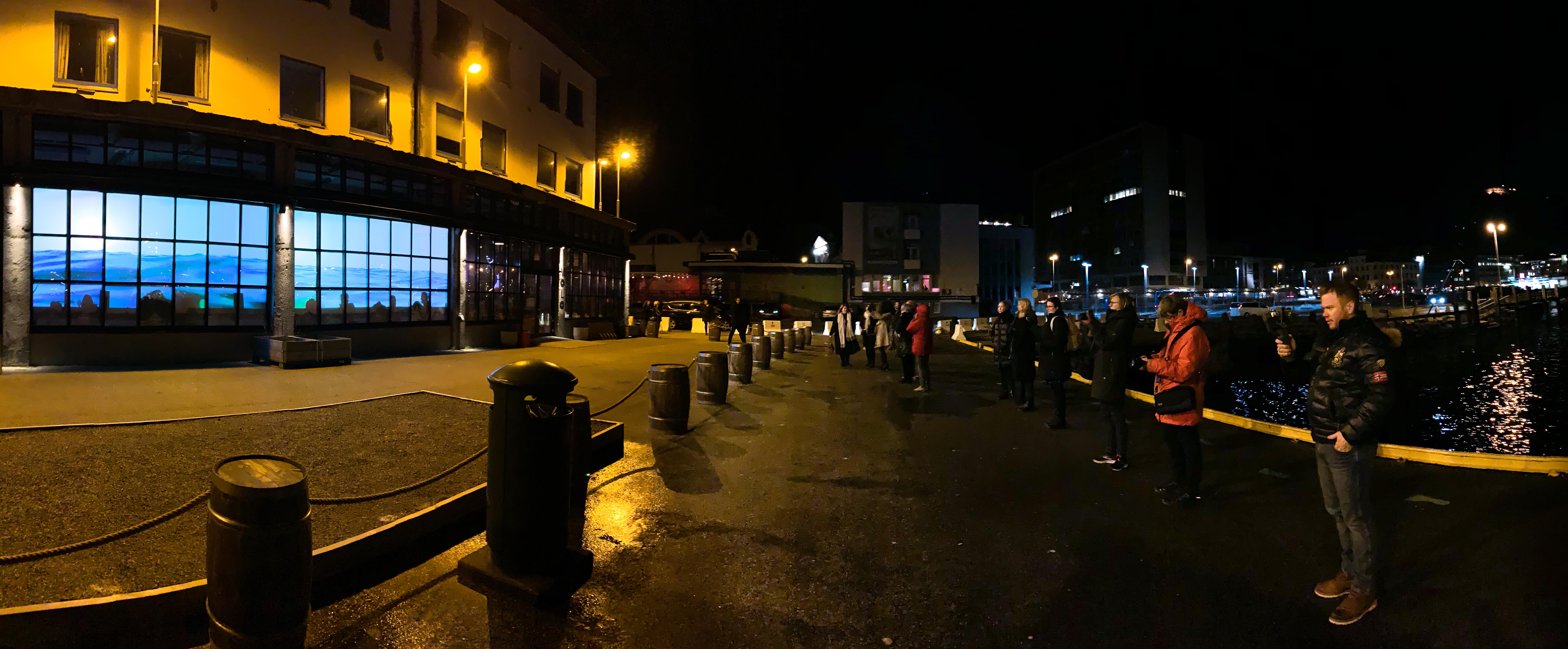 Panoramic photograph with Water Will Be Here - a video installation projected into the windows of a bar showing water ebbing and flowing slowly - on the left and several spectators outside of the bar on the right, with a body of water behind them