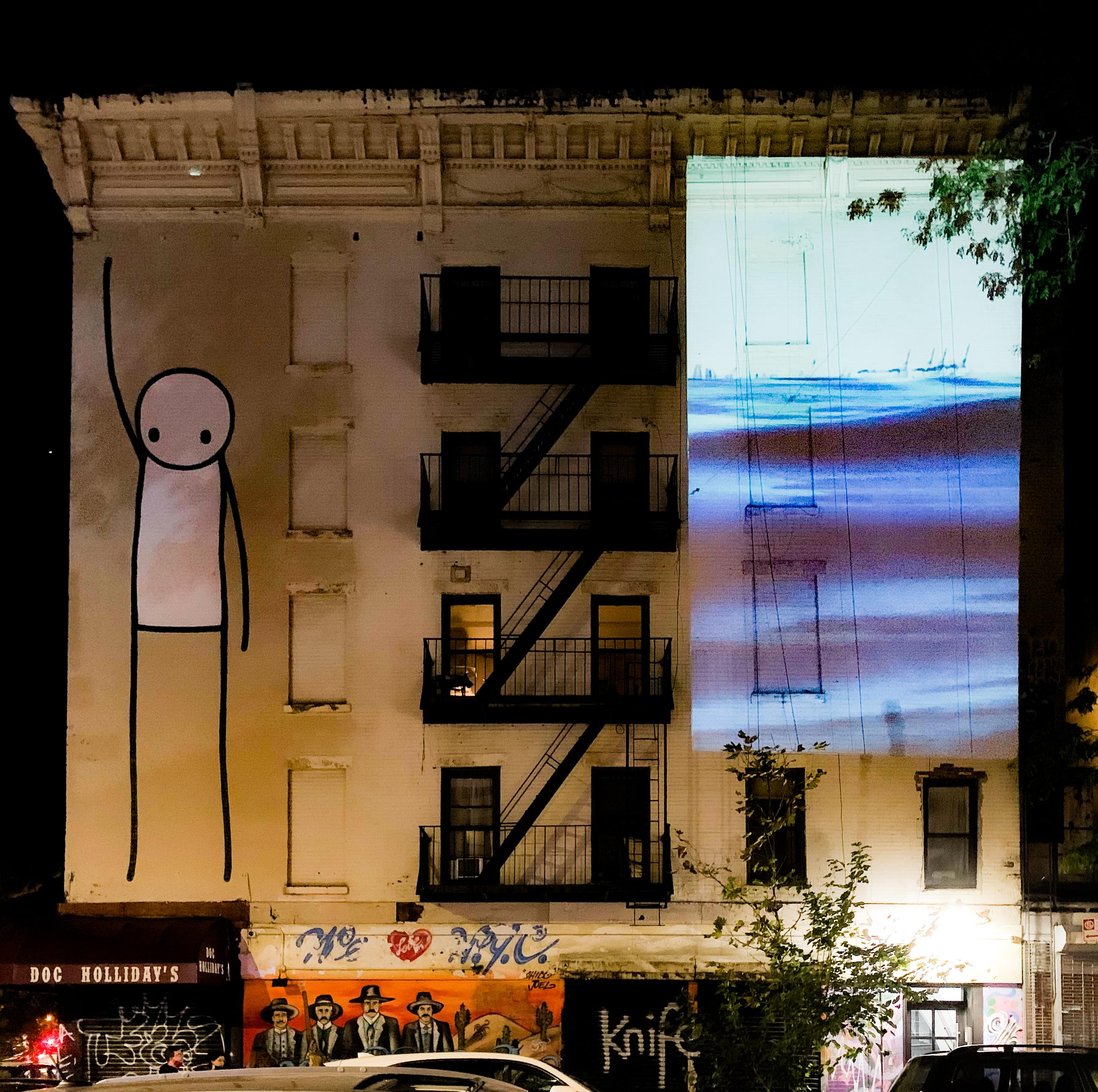 Photograph of a white building in New York City which has a 35-foot-tall graffiti drawing of a stick figure reaching towards the sky on the left and Water Will Be Here projected, equally as tall, on the right