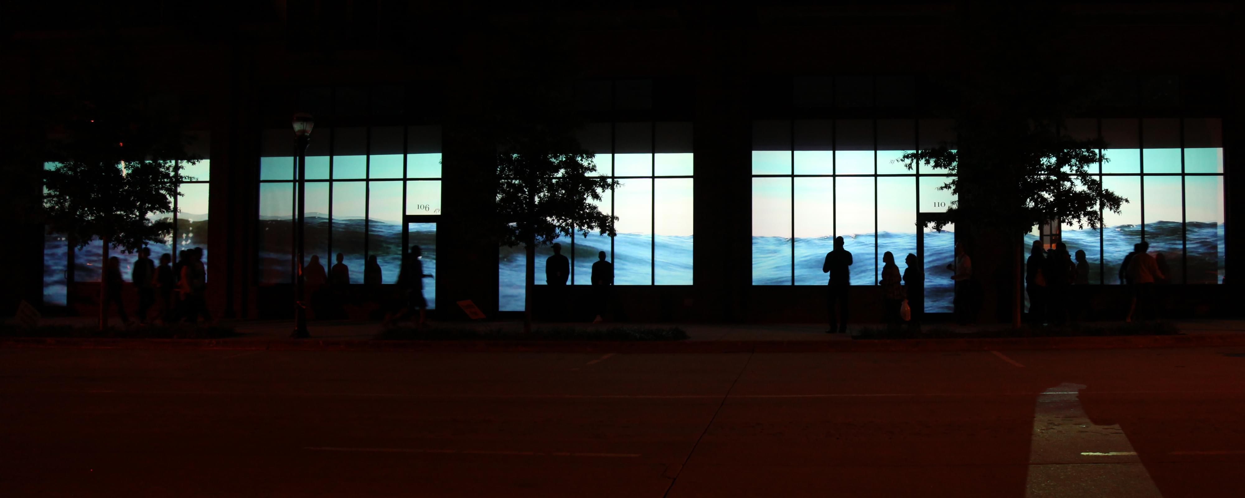 Nightime photograph of a several storefronts with a projection of water (from the sea) in their windows. Water level is contiguous across many storefronts. Inside the storefronts are silhouettes of people interacting with the video installation.