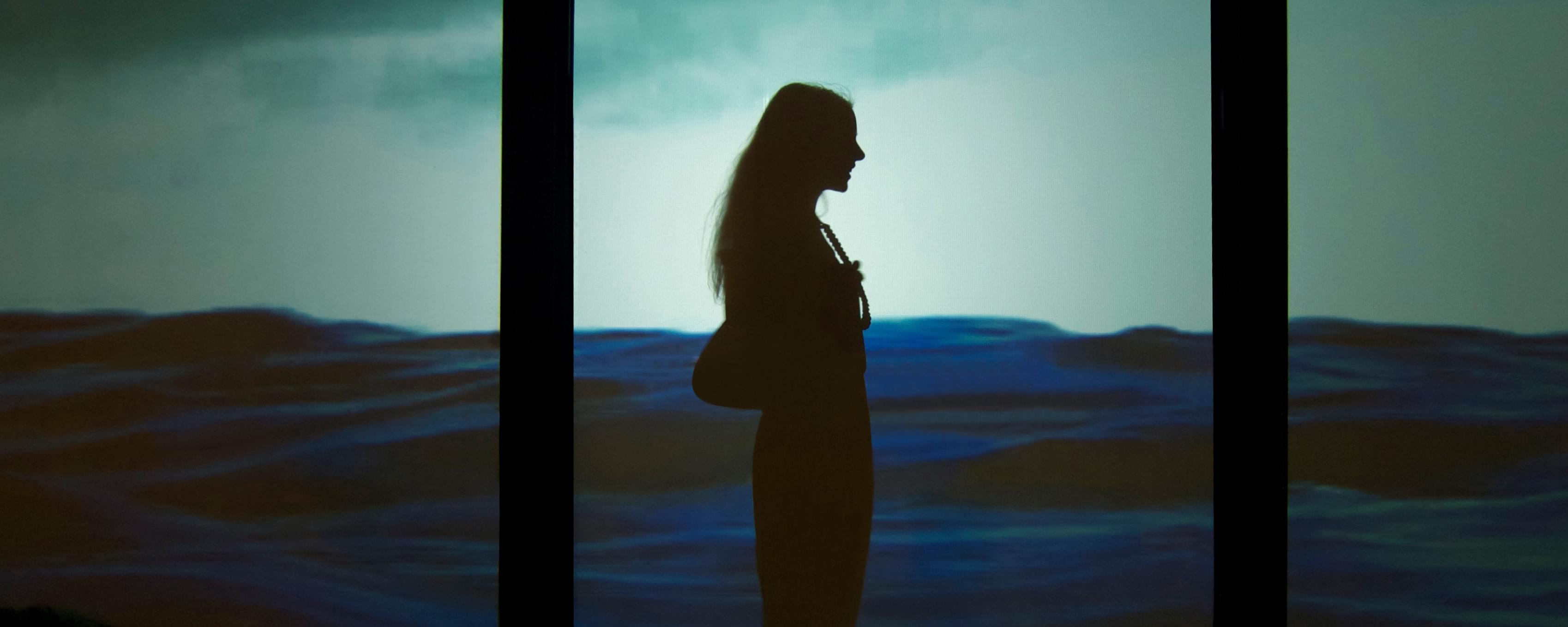 Photograph of a women's silhouette is shown in front of a projection of a body of water that is up to her chest; a blue sky is behind her