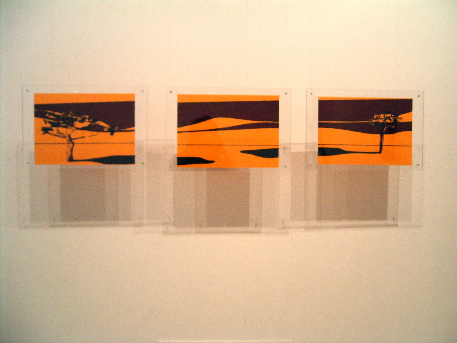 Photograph of three artworks, encased in transparent plexiglass, hanging from the ceiling against a white wall. Each artwork contains an element of a triptych, which taken together form a golden/yellow-ish abstract landscape with one tree on each side. The hanging artworks cast a complex array of shadows onto the wall behind.
