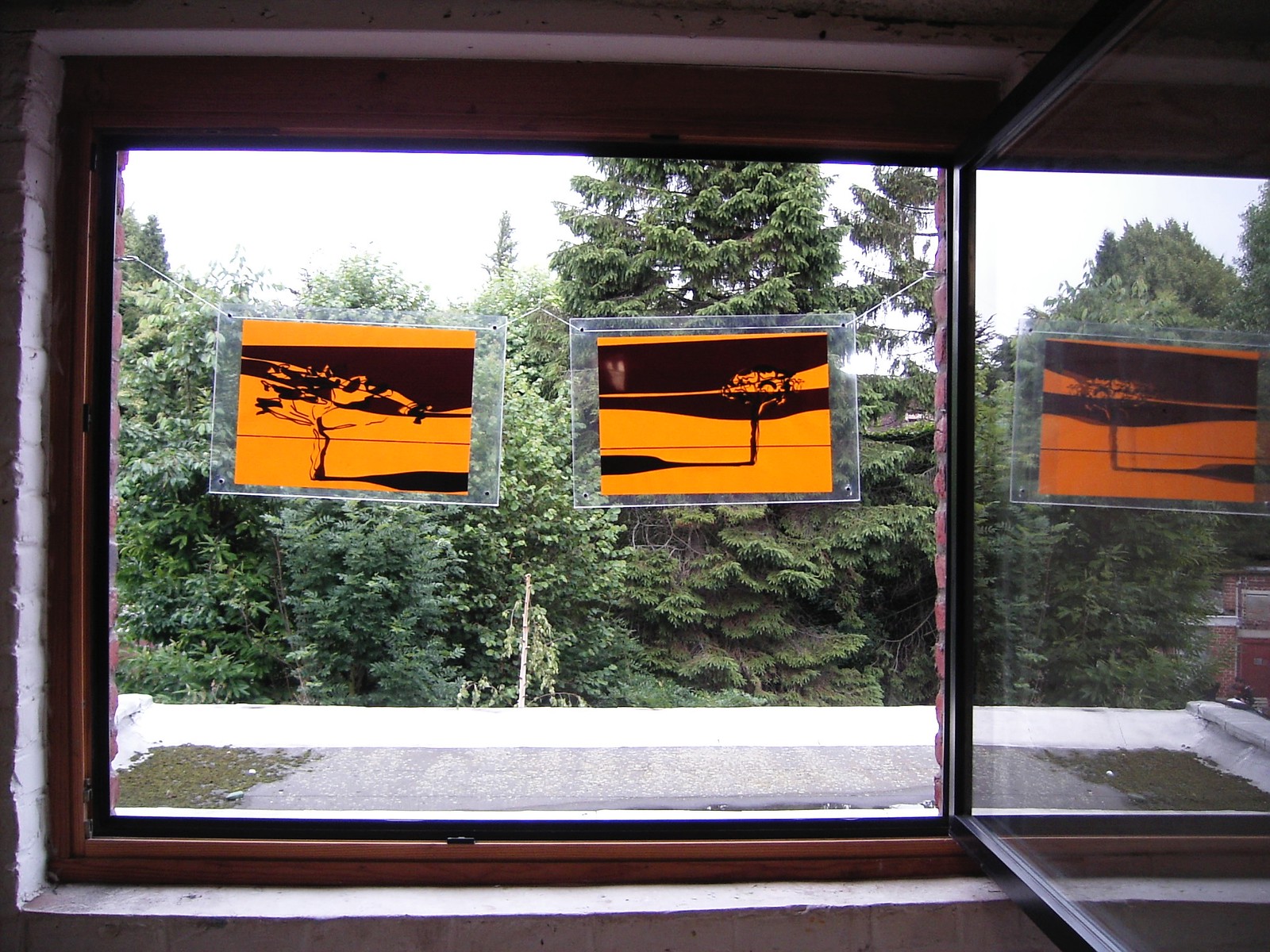 Photograph of two artworks, encased in transparent plexiglass, hanging in a large open window. Each artwork contains an element of a diptych, which taken together form a golden/yellow-ish abstract landscape with one tree on each side. Several trees are visible behind the hanging artworks in the open window.