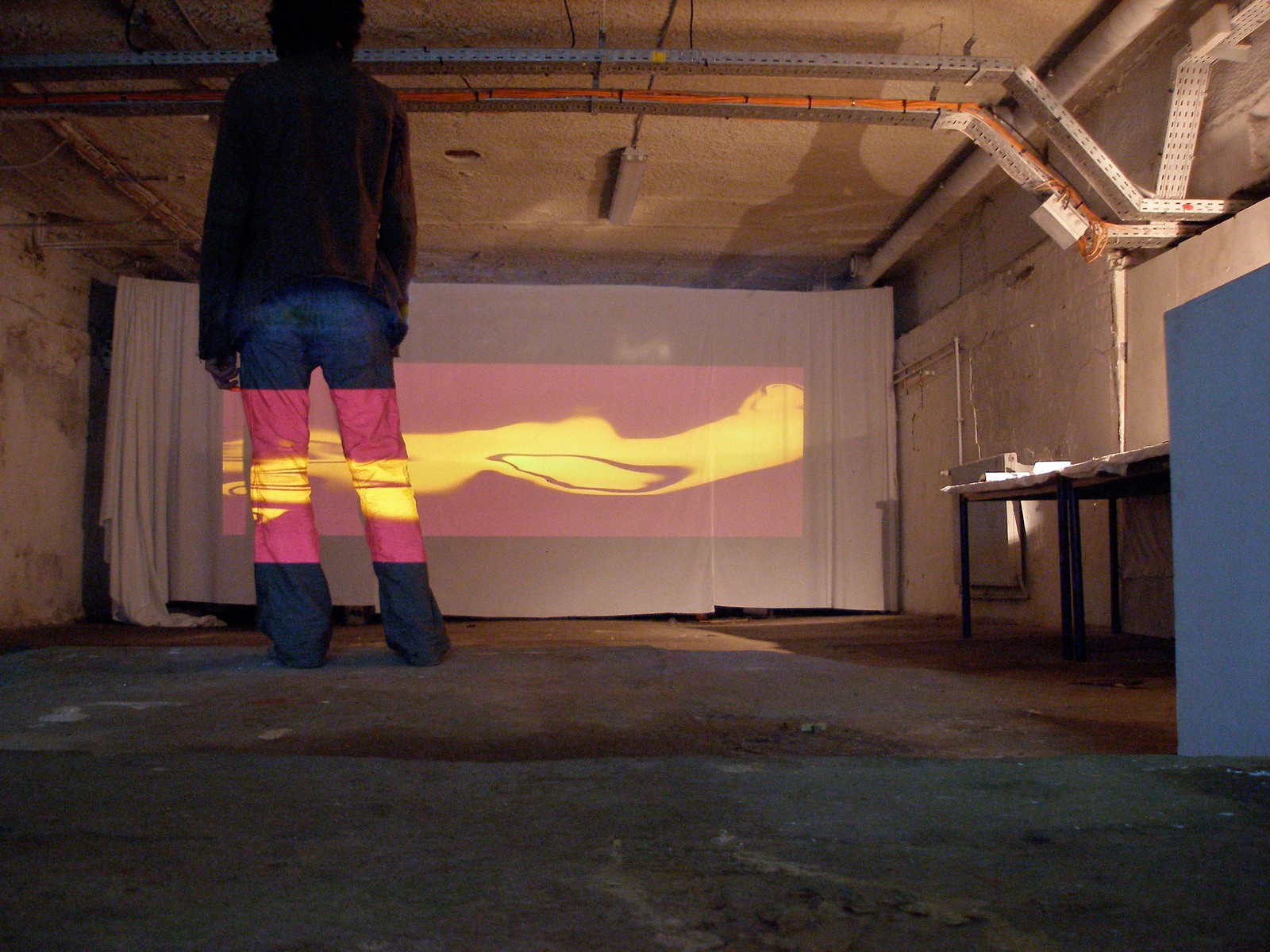 Photograph of the back of a person, wearing blue jeans and a dark jacket, with a projection of abstract burgundy and yellow organically shaped forms that are also projected onto a large screen at the end of an industrial-looking space.