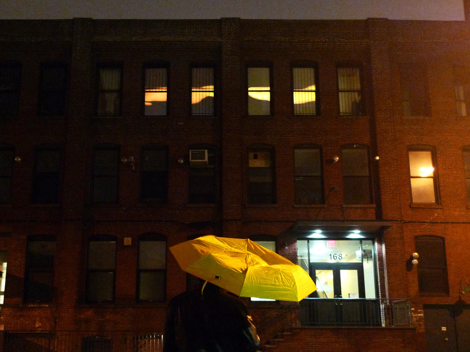 Nighttime photograph of a large, three-story industrial brick building. In the top floor, four center windows display a projection of an abstract, fluid yellow band. In the foreground is a person standing with a yellow umbrella.